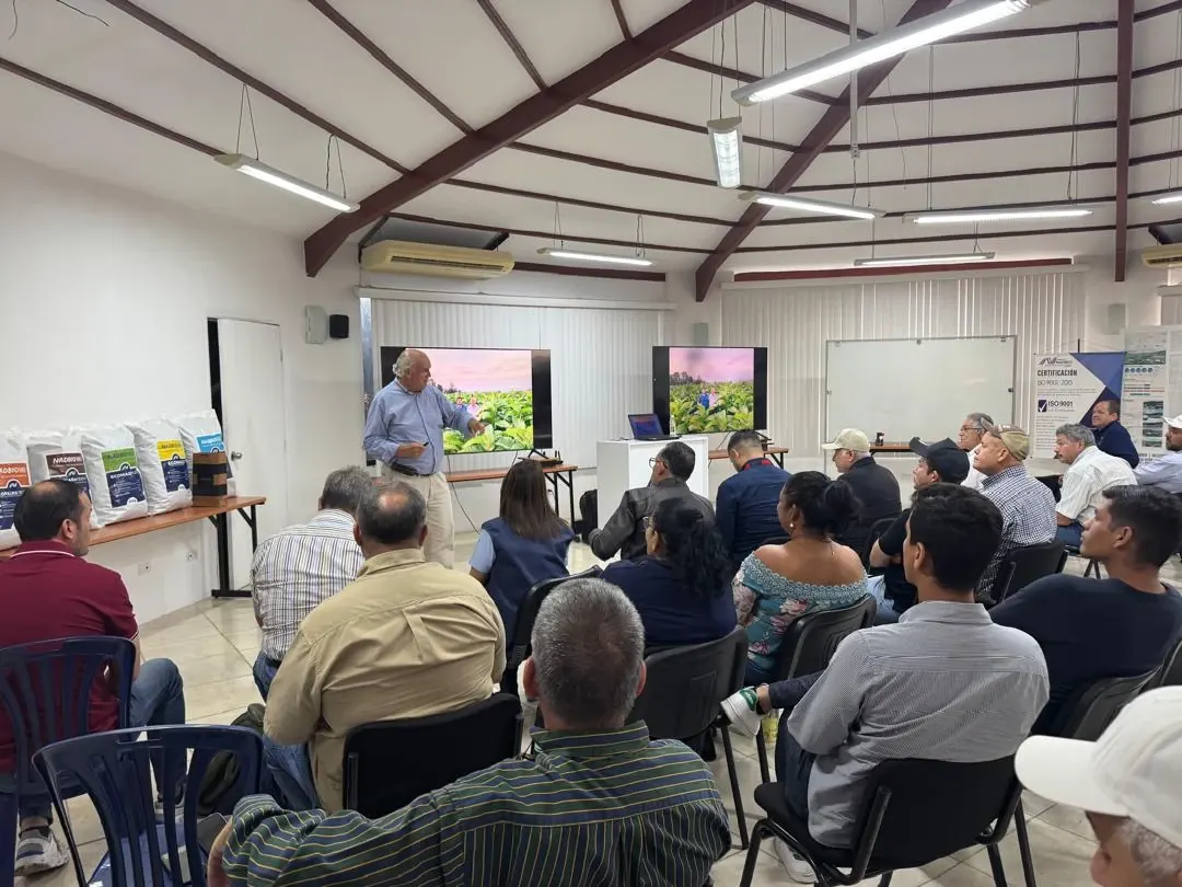 Charla técnica en un salón: un ponente explica frente a dos pantallas con fotos de un cultivo, ante productores sentados.